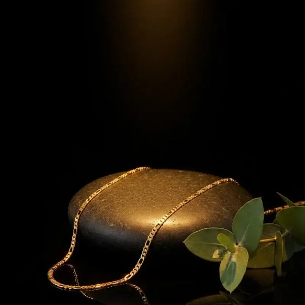 18kt gold titanium steel Figaro link chain resting on a smooth black stone with greenery.